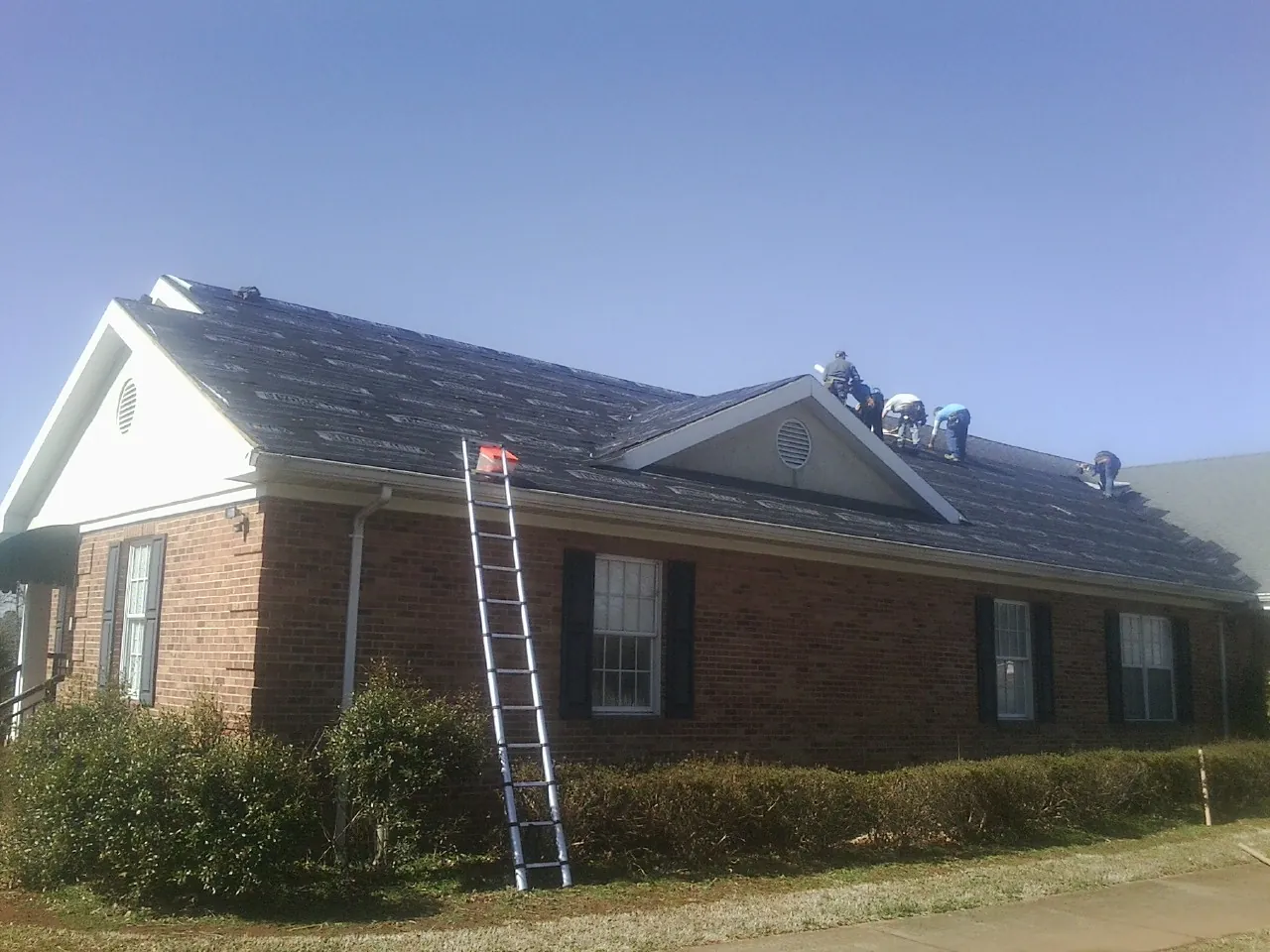 Crew tearing shingles off a brick ranch roof with a ladder set — Metal Roof Repair work by our crew in George West